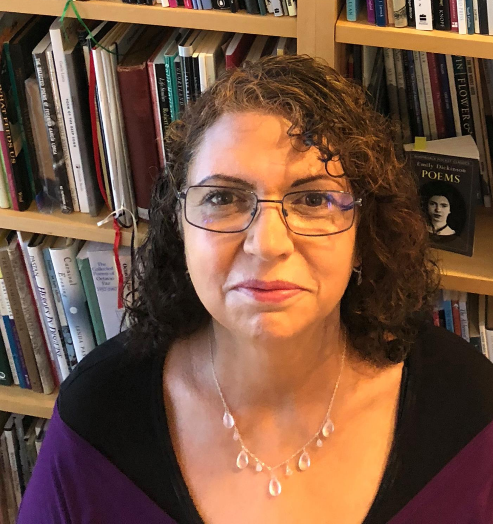 A white middle aged female wearing glasses and a silver necklacesits in front of a book case. 