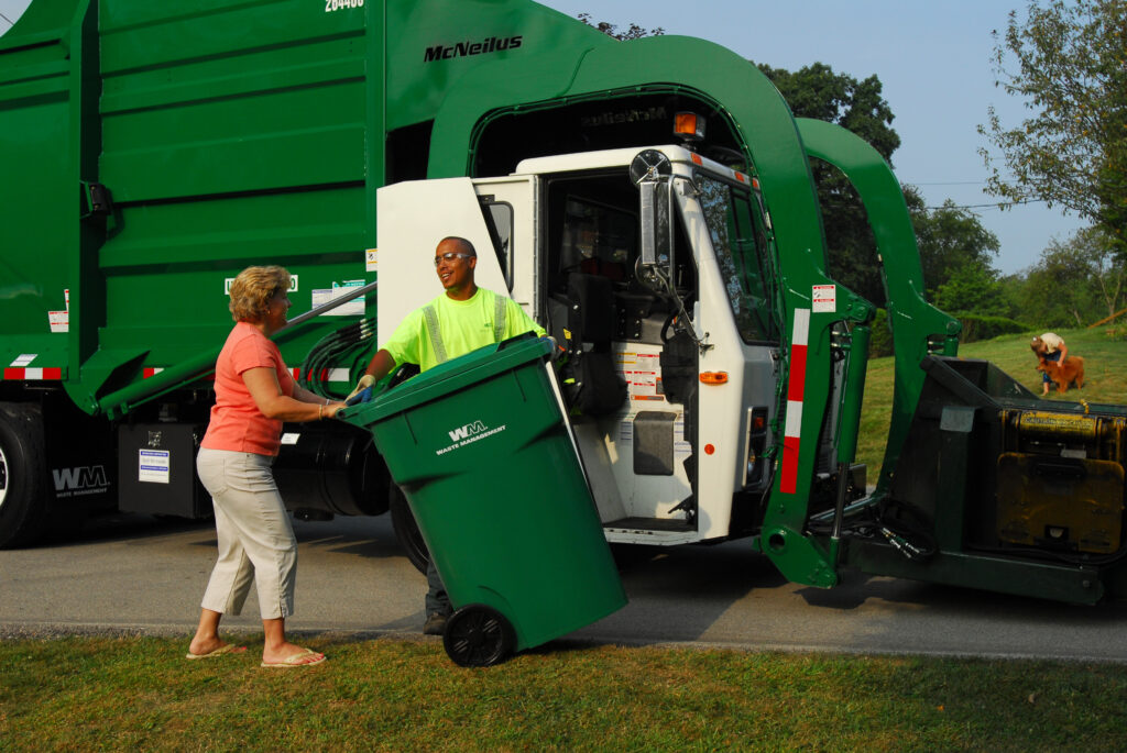 A Waste Management truck is parked next to the curb, the Waste Management driver speaking to a female customer who is posed to bring in her container.