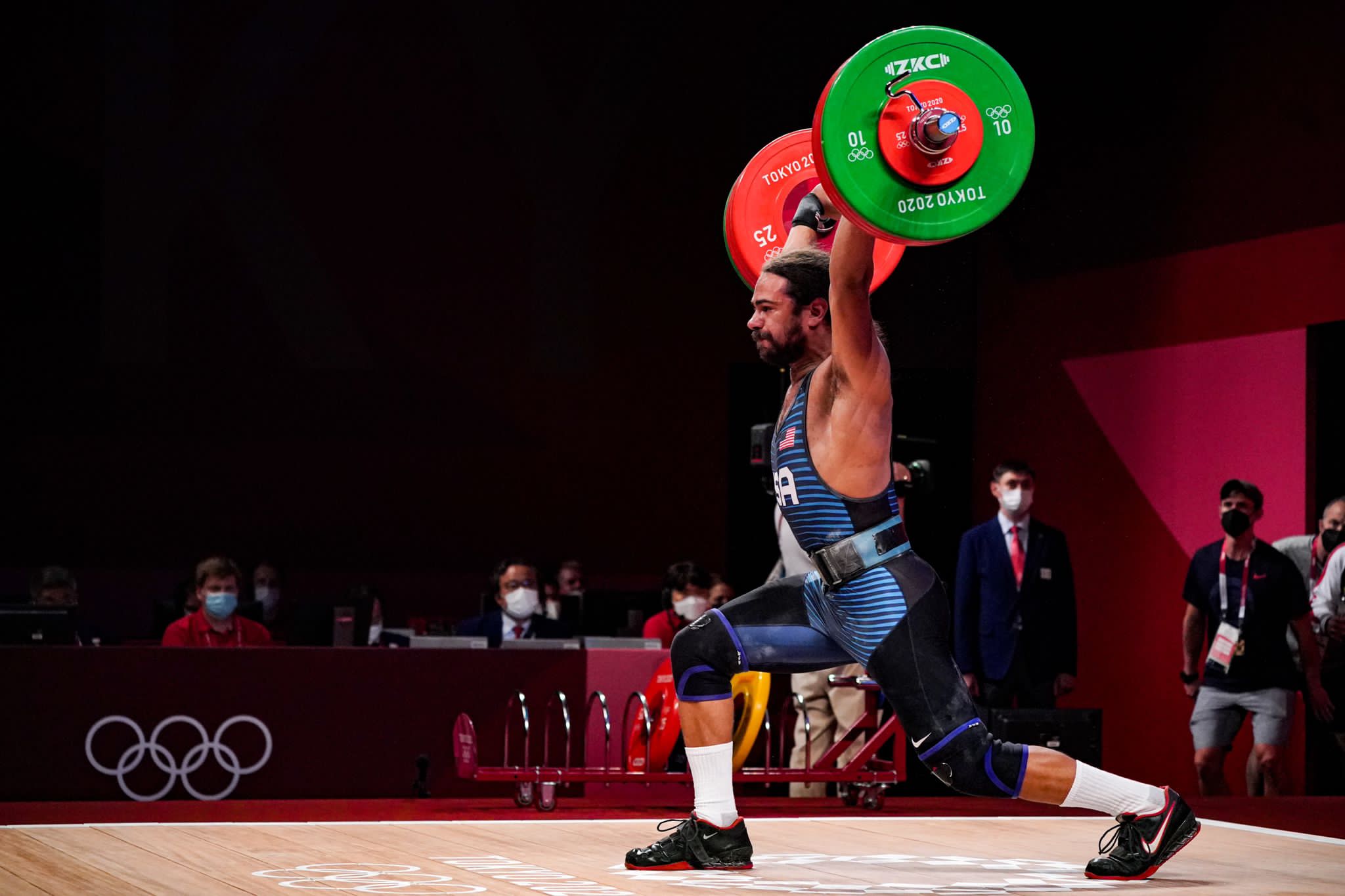 Weightlifter Harrison Maurus hoists a heavy barbell over his head, one leg bent slightly at the knee and the other behind him in a brace position.