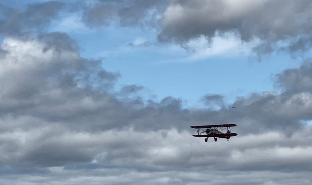 A restored Boeing Stearman biplane flies through ablue sky with gray clouds.