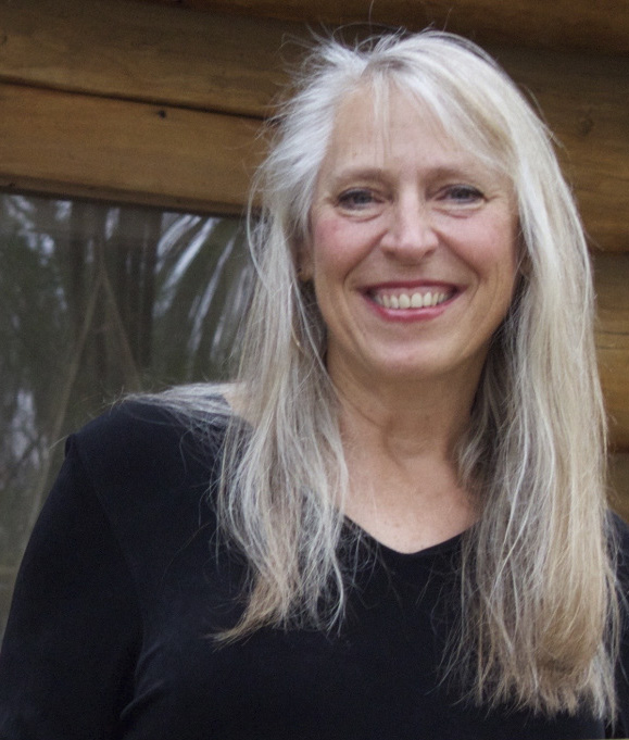 Cindy Ross, a middle-aged white woman with long light blonde hair, smiles widely at the camera white standing in front of a window.