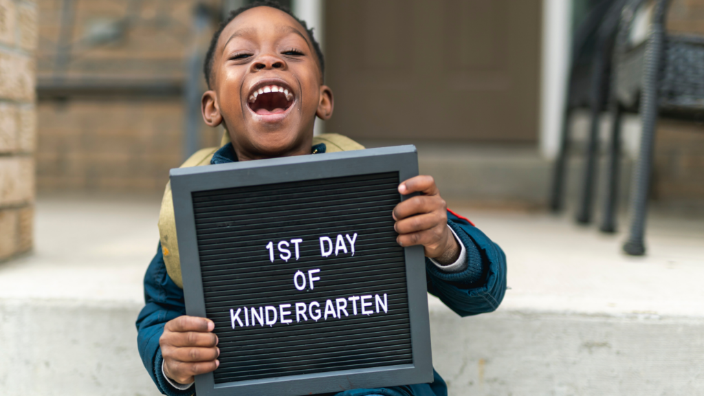 a laughing child holds a sign that says "1st day of kindergarten"
