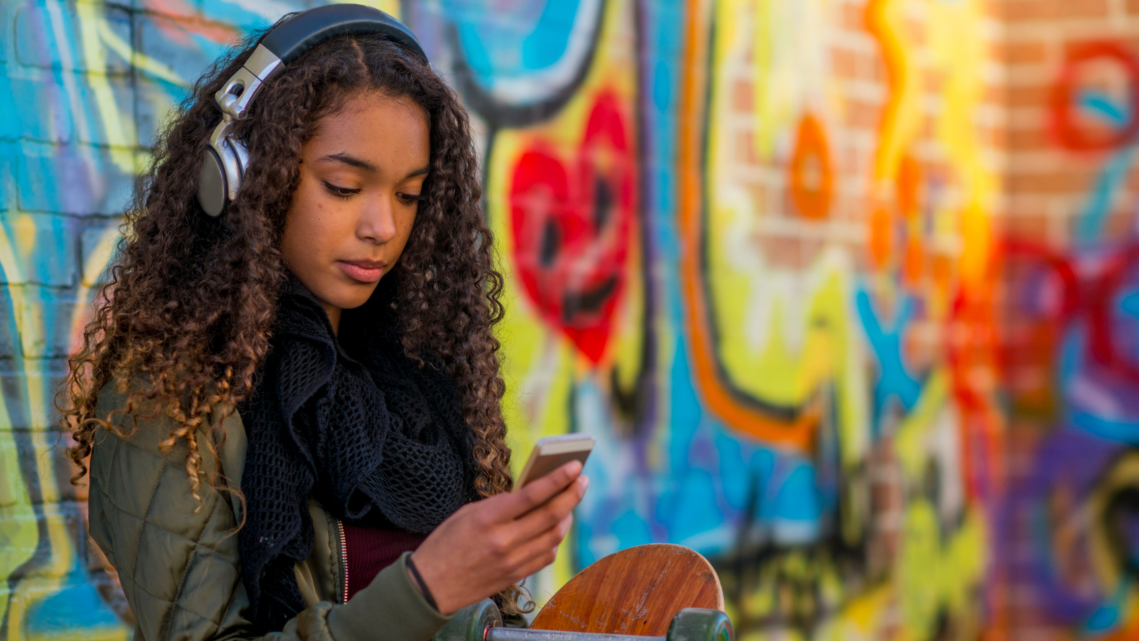 A young female looks at her phone as she listens to music on headphones, she leans against a vibrantly colored mural on a brick wall
