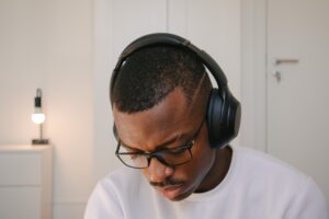A Black male wearing glasses and a white shirt wearing large headphones looks downward