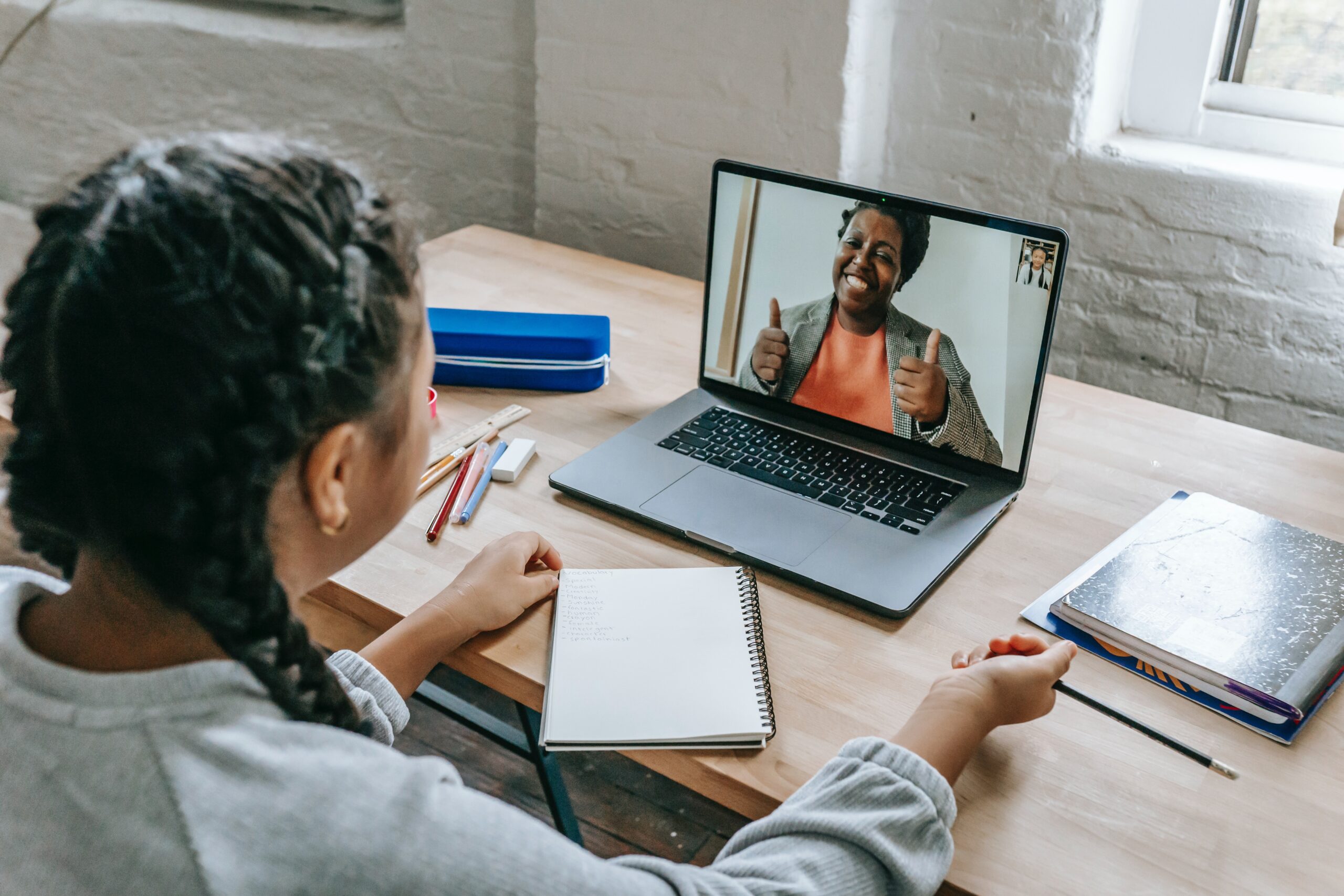 a young girl with braids sits before a laptop computer, a Black female on the screen smiling and giving her a thumbs up.