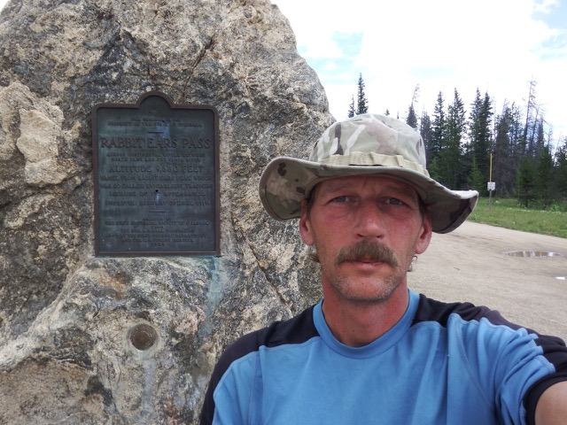 Shawn Murphy takes a selfie on a trail next to a marker embedded in a large boulder. He wears a floppy fisherman's bucket hat and a blue t-shirt
