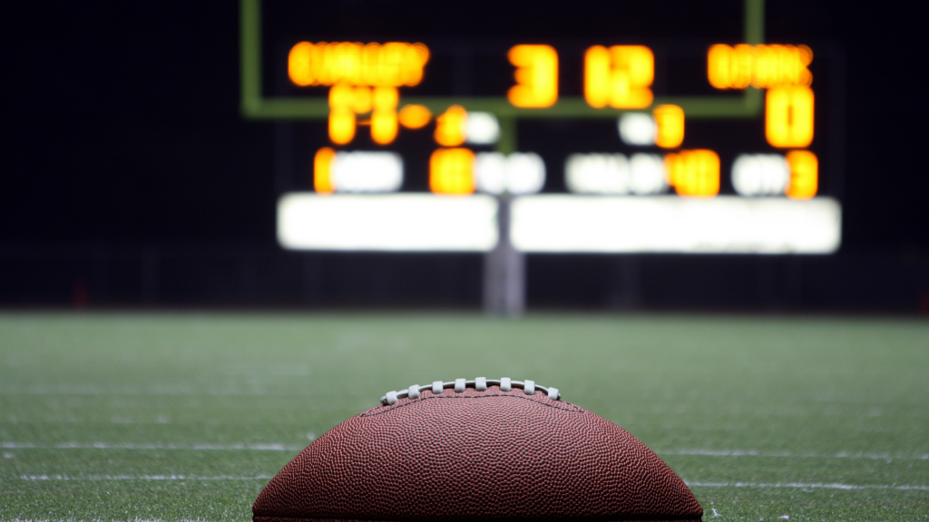 A football in the foreground of a blurred football field and lit up scoreboard