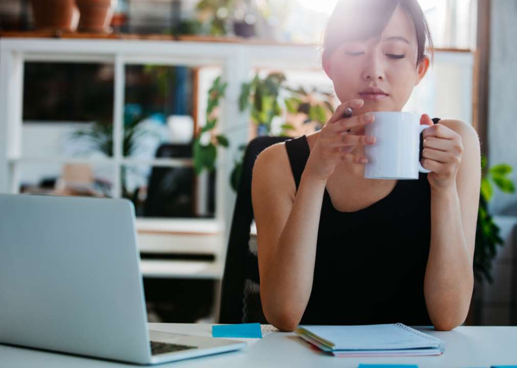 An Asian female holding a coffee mug up toward her face sits at a desk with a laptop and pad of paper in front of her.