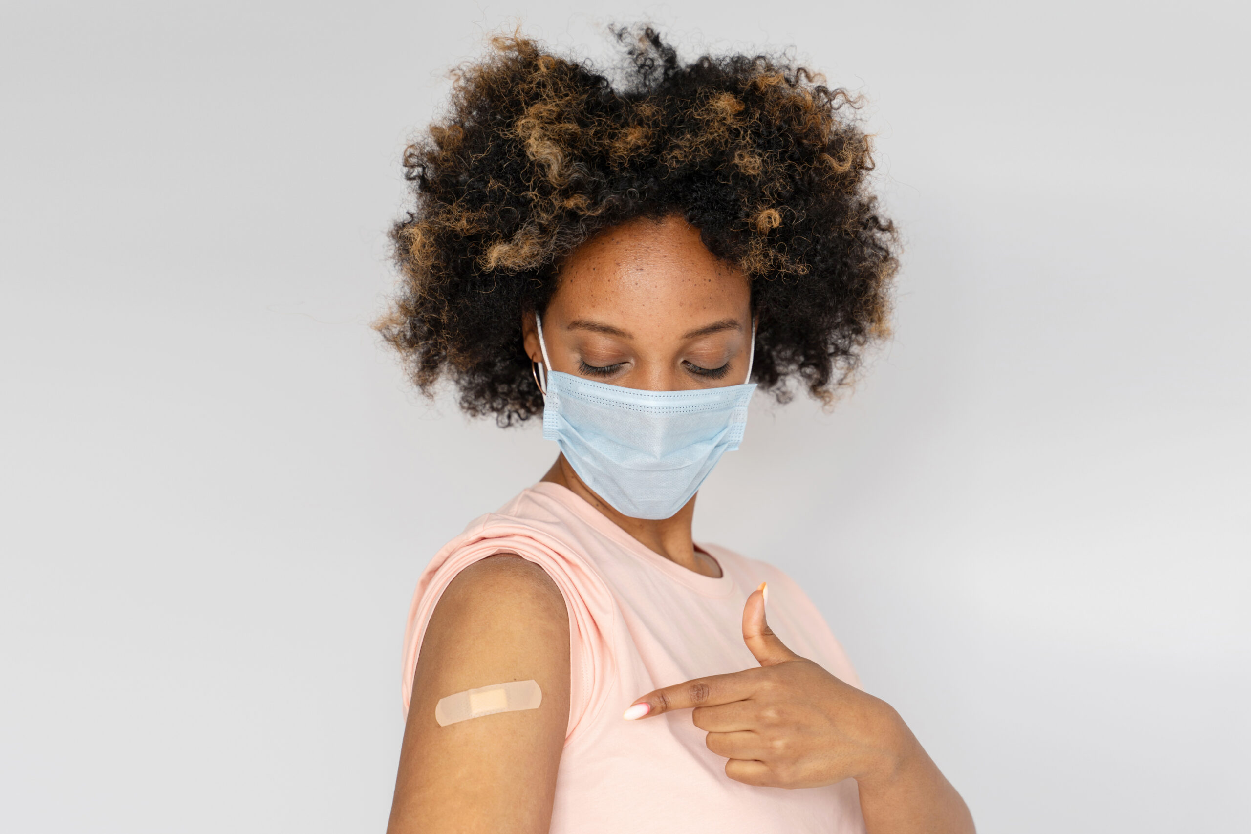 Young african american woman in medical mask looking at her arm with band aid after receiving first dose of coronavirus COVID-19 vaccine injection