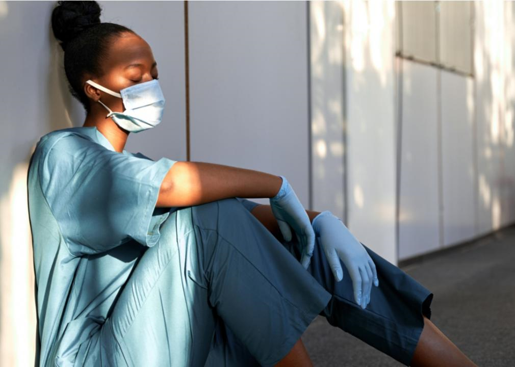 A female medical professional sits on the ground leaning against a wall, visibly tired