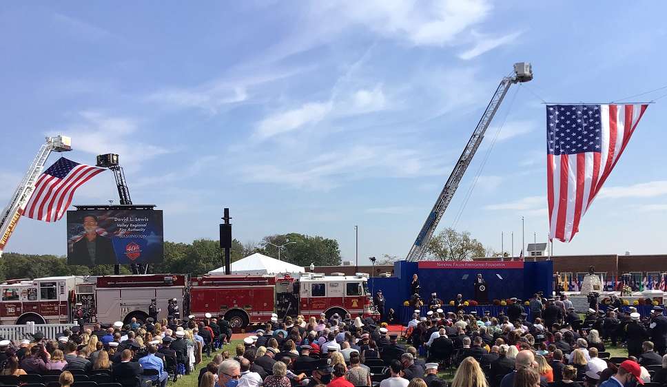 Two large American flags hang from firetruck ladders, a crowd of people listen to names announced from an honor roll of fallen firefighters. Dave Lewis' name and photo are on a jumbo screen at the front of the crowd