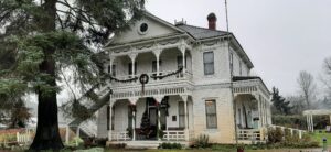 A historic mansion decorated in victorian style holiday decor. In front of the two story white 1894 farmhouse mansion is a large tree.