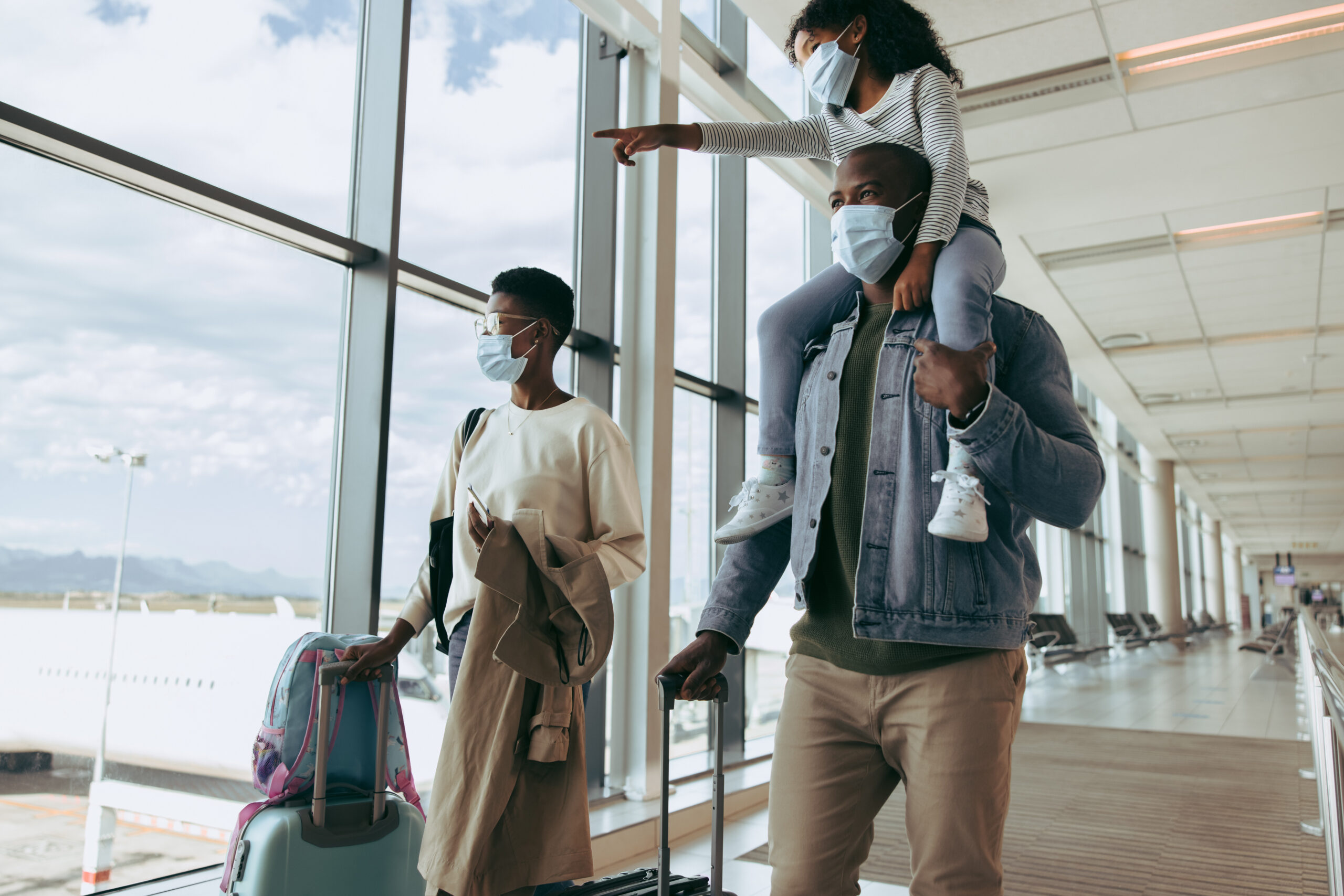 Kid girl on shoulder of man showing airplanes while walking at airport. Family wearing face masks going for boarding airplane.