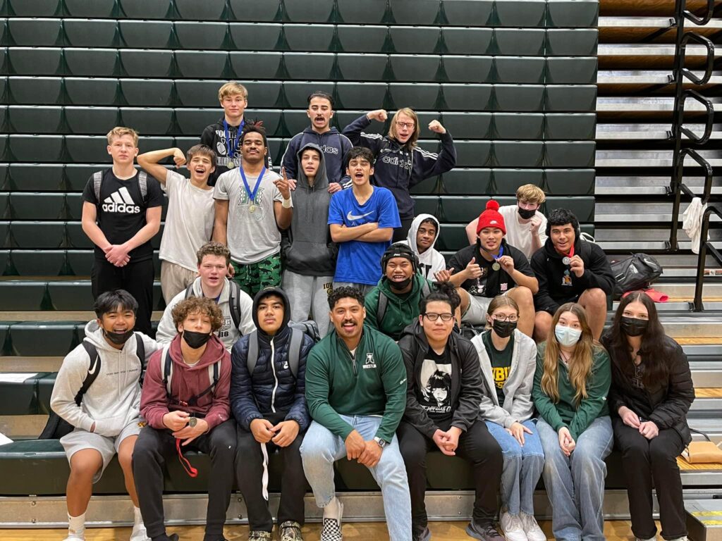 Auburn High School students and wrestling coach sit on bleachers for a group photo. Some wrestlers wear medals around their neck. 6 students wear masks.