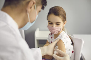 Brave girl in face mask looking at needle in doctor's hands while getting a flu shot at clinic or hospital. Medical worker injecting little kid with Covid-19 vaccine. Immunization for children concept