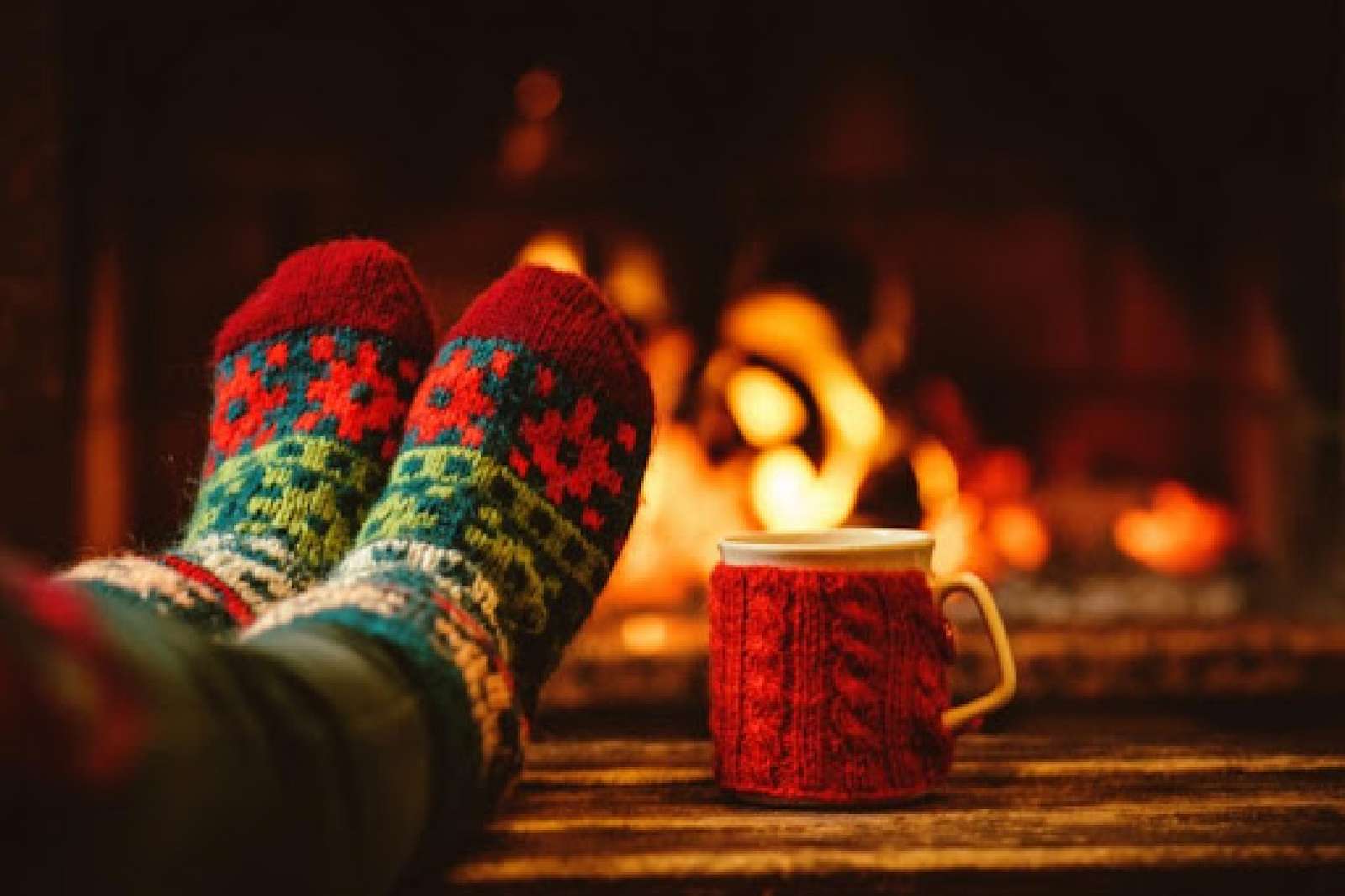 A pair of feet wearing colorful holiday socks rest on a table next to a mug wrapped in a knitted koozi. A crackling fire is blurred in the background.