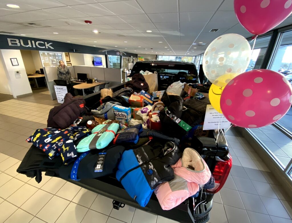 A black pick up truck with balloons on it inside a dealer showroom.. The bed of the truck is filled with donated clothing and food items.