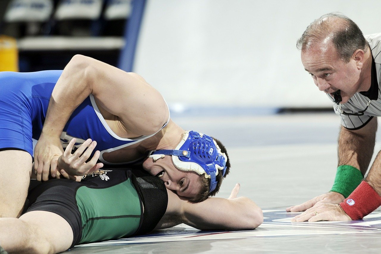 Two high school wresters in a match, one pinning the other. A ref leans in closely with a whistle in his mouth