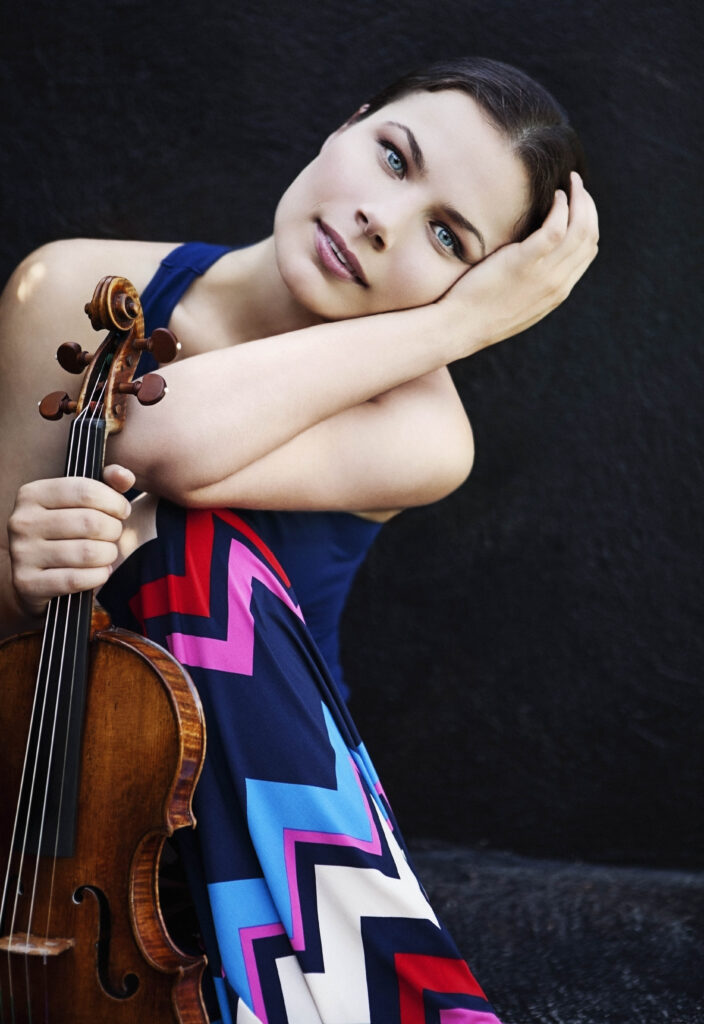A brunette woman holding a violin at the top of its neck looks toward the camera, her cheek resting against her wrist