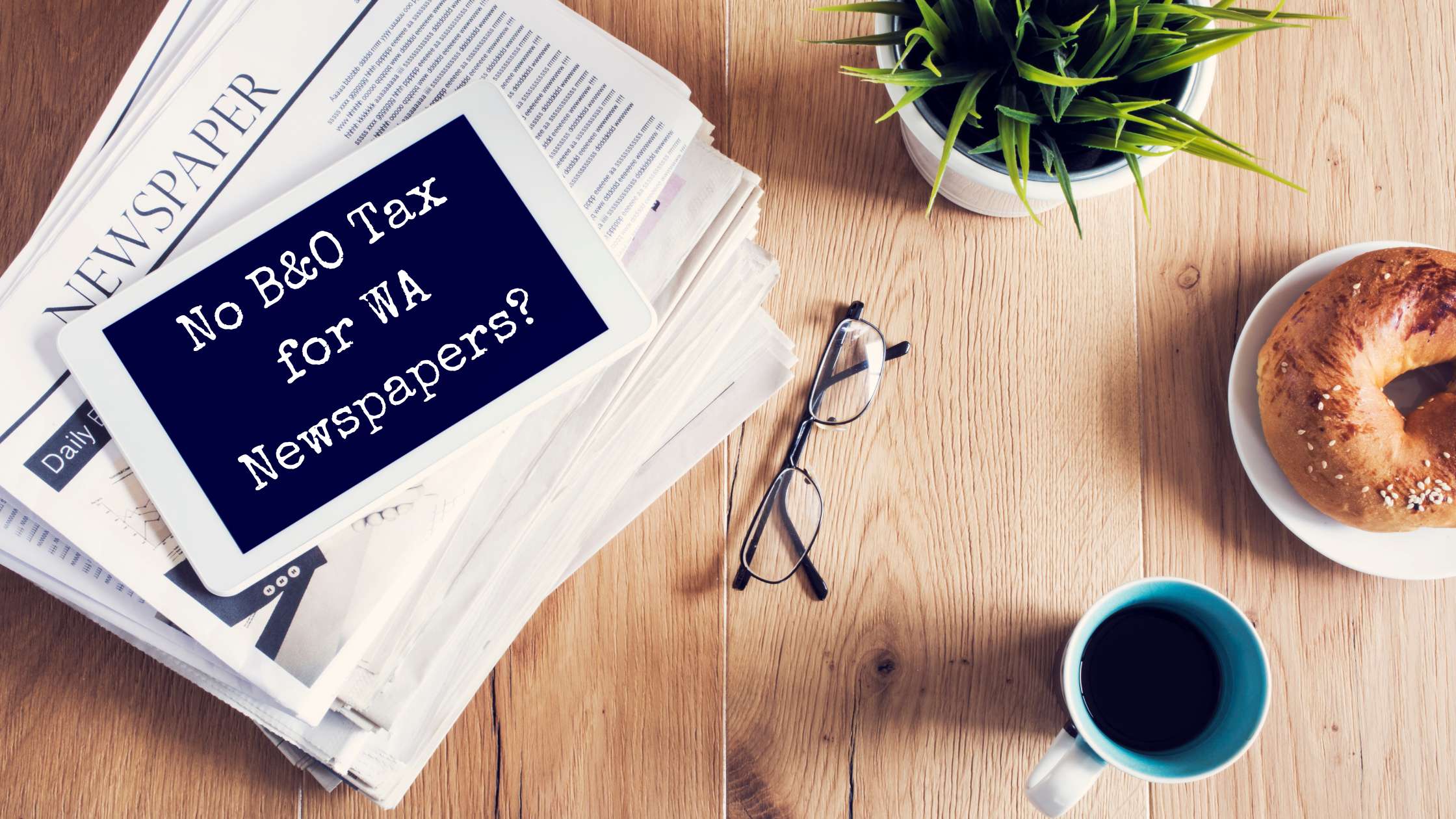 A tablet ontop of a stack of newspapers, a plant, coffee cup, bagel on a plate, and pair of glasses lay across a desk.
