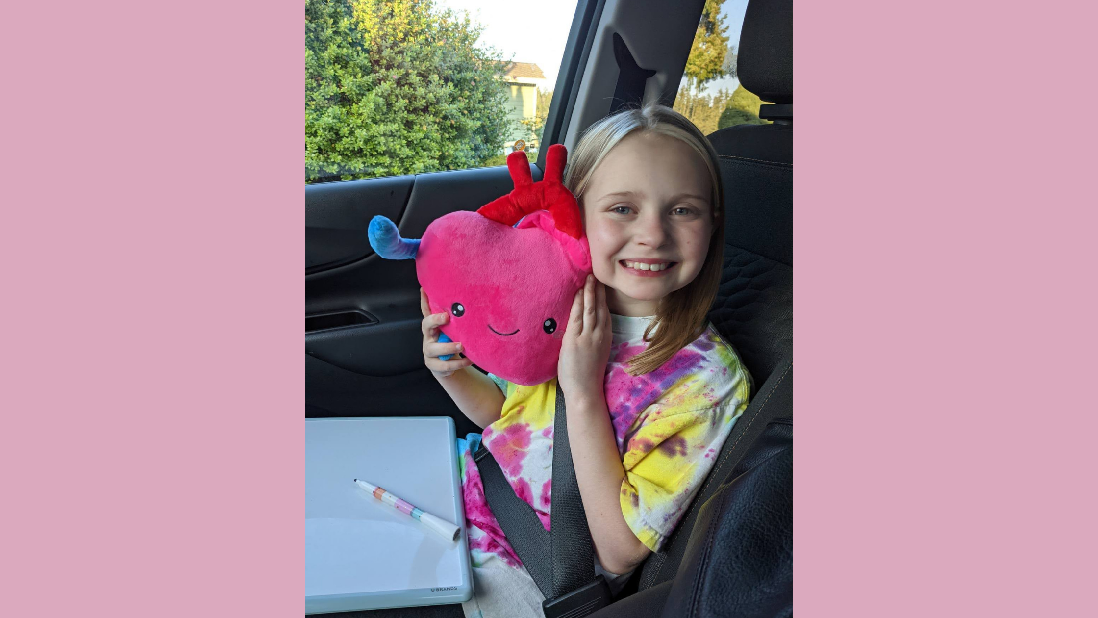 Sitting in a car, a young blonde girl smiles as she holds a stuffed heart up to her cheek.