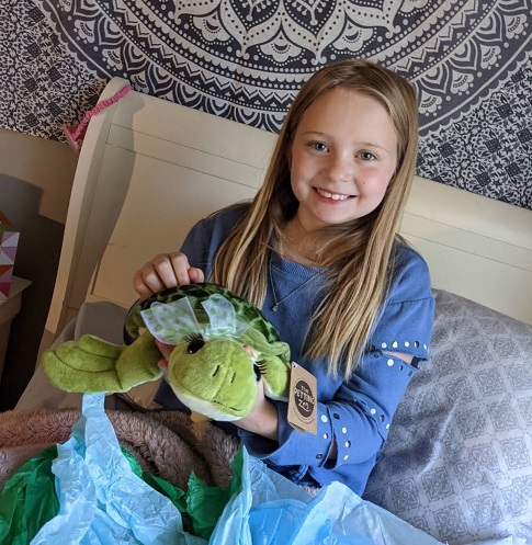 A young blonde girl smiles as she shows a stuffed turtle to the camer