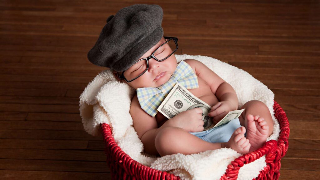 A baby in a red basket sleeps holding a $100. The baby wears glasses, a bowtie and a cute hat