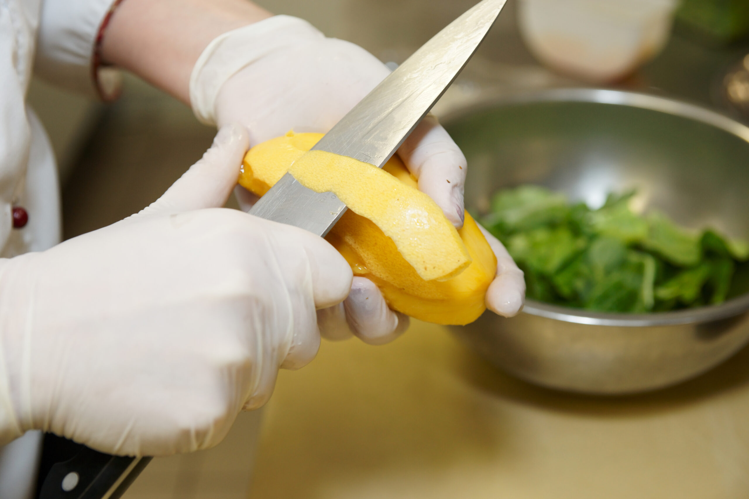 A pair of hands wearing white latex gloves uses a chef's knife to cut a yellow mango. A bowl of salad is blurred in the background.