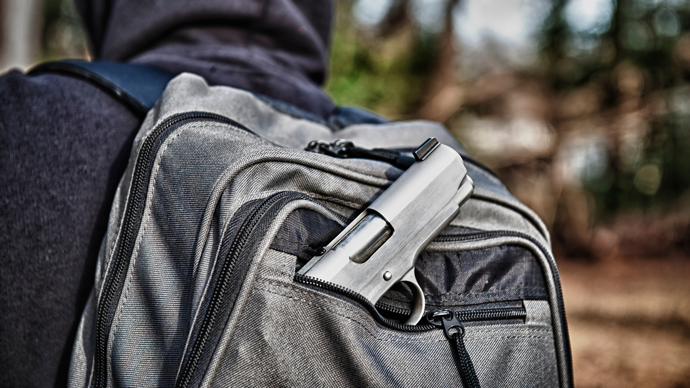 A black handgun rests part way in the front pocket of a black backpack on the back of a hoodie wearing individual