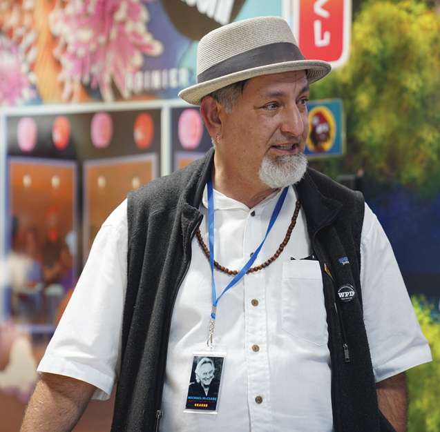 A photo of Paul Nelson, an older male with olive skin and a white goatee wearing a fedora, white shirt and open black fleece vest looks to the side.
