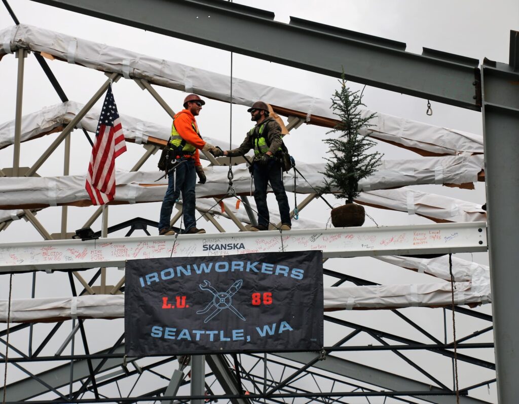 Two ironworker construction workers stand on a metal beam in a school structure.