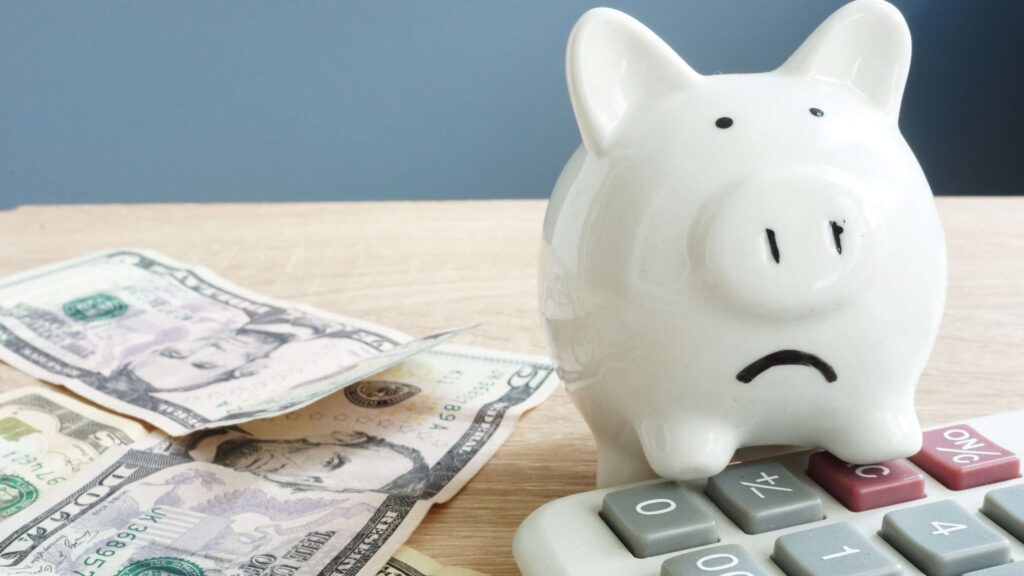 Struggling with financial anxiety a piggy bank frowns, sitting on the edge of a calculator with a few dollars tossed next to it. bank frowns as it struggles with f