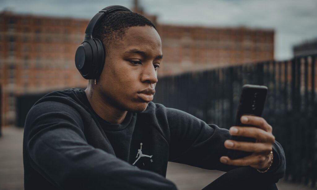 A young adult Black male wearing headphones looks at his phone as he listens to music.