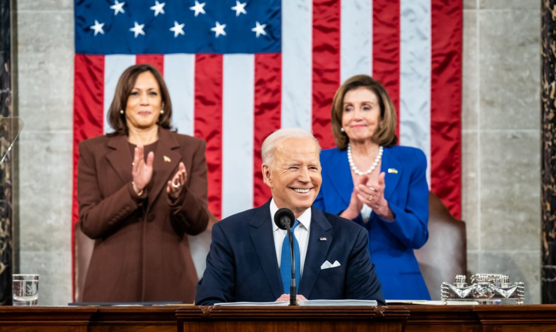 President Biden sits before a microphone, VP Harriw and Speaker Pelosi standing behind him applauding. He is preparing to give his first State of the Union address