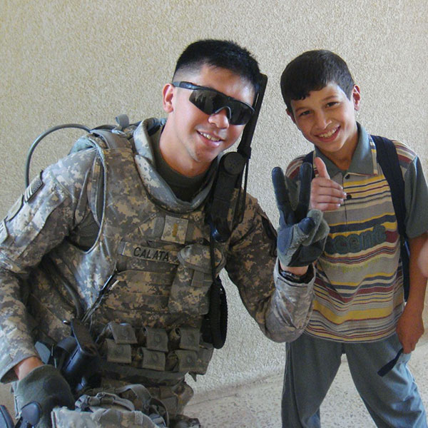 Deputy Dom Calata dressed in his fatigues gives the peace sign standing next to a young boy wearing a backpack and smiling