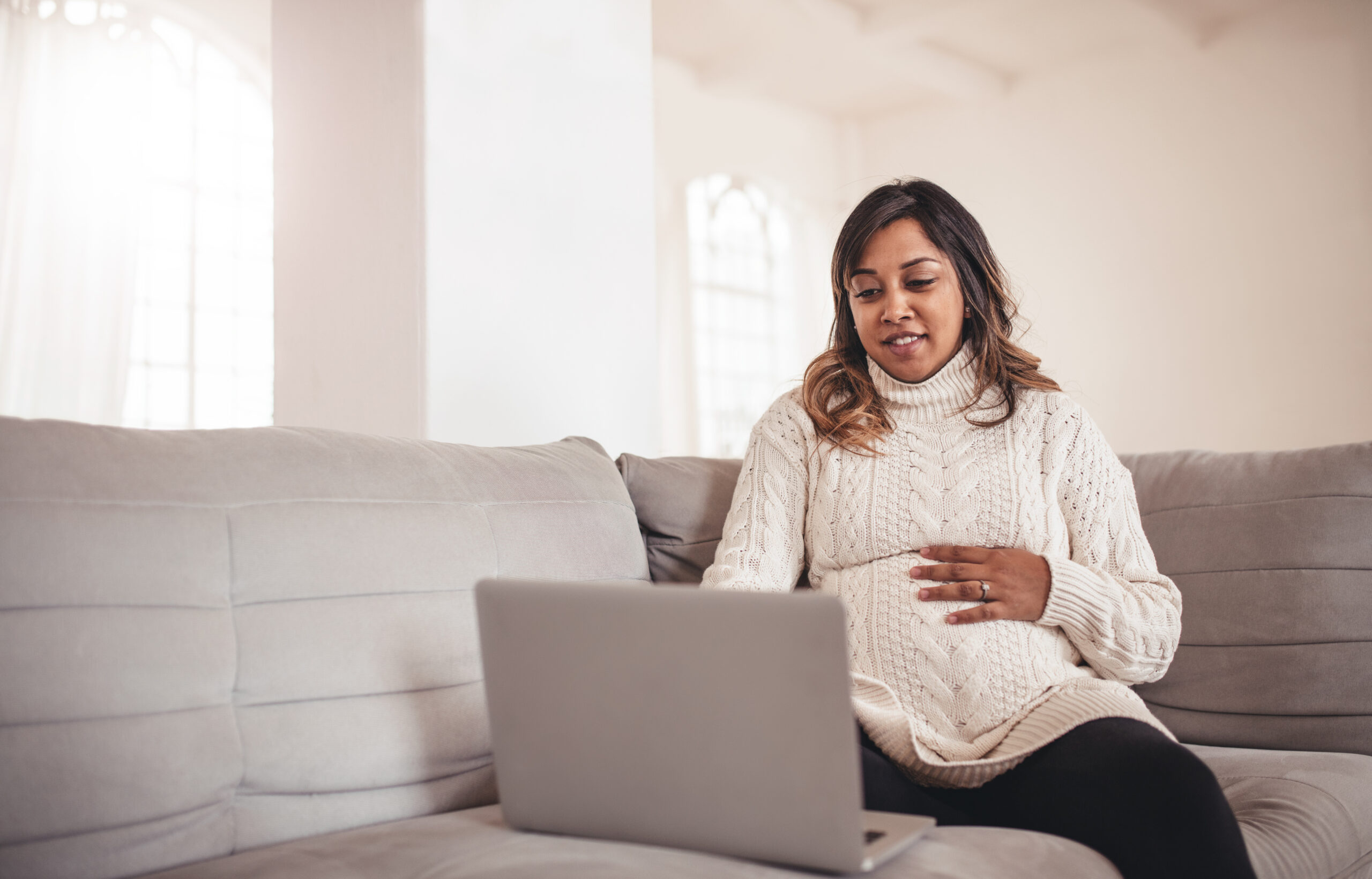 Portrait of beautiful young pregnant woman with laptop sitting on sofa. African woman expecting a baby sitting on couch and using laptop at home.