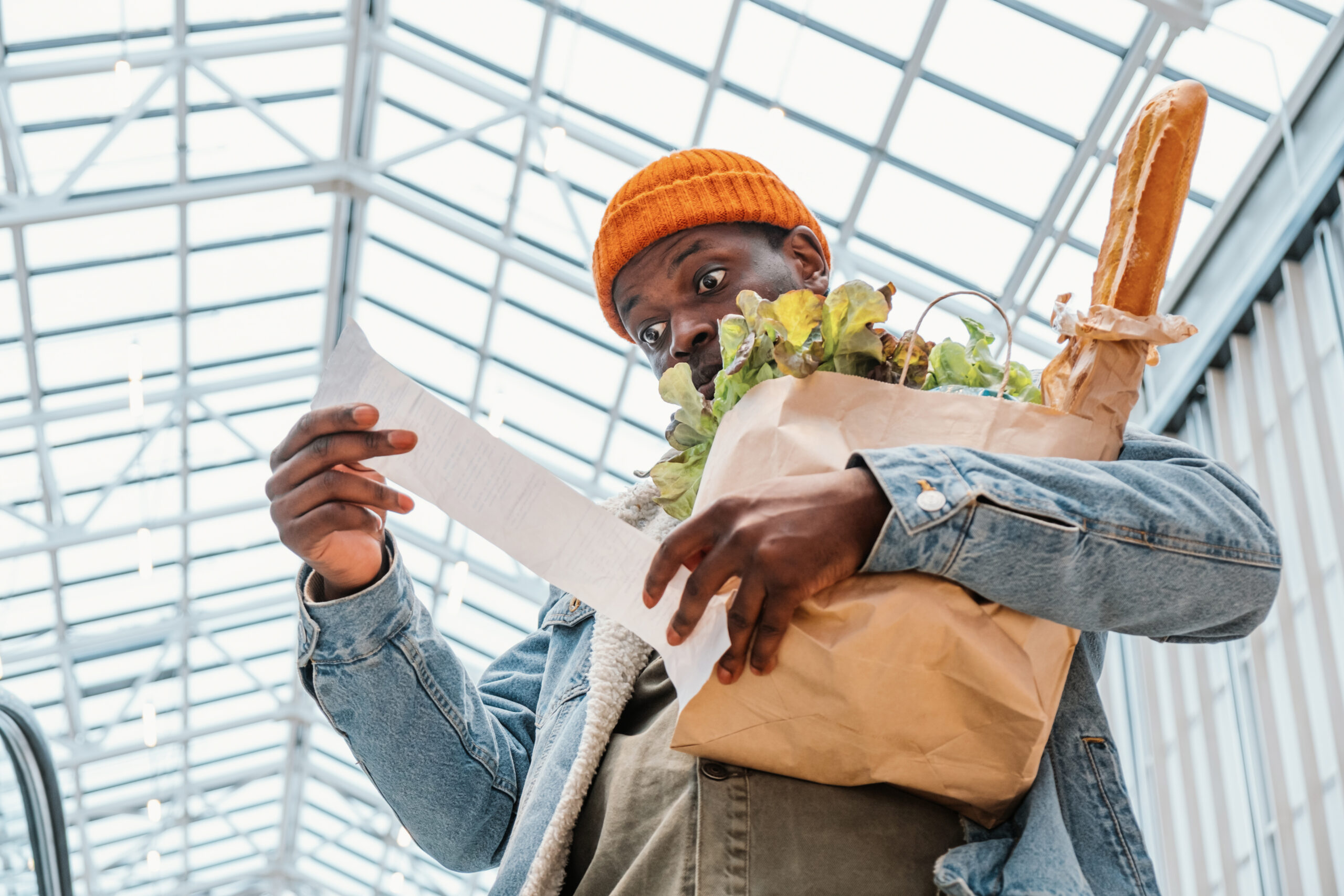 Surprised African-American man in denim jacket looks at receipt total in sales check holding paper bag with products in mall