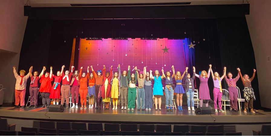 Students in bright colored costumes stand across the front of the stage with their hands lifted in a final curtain call.