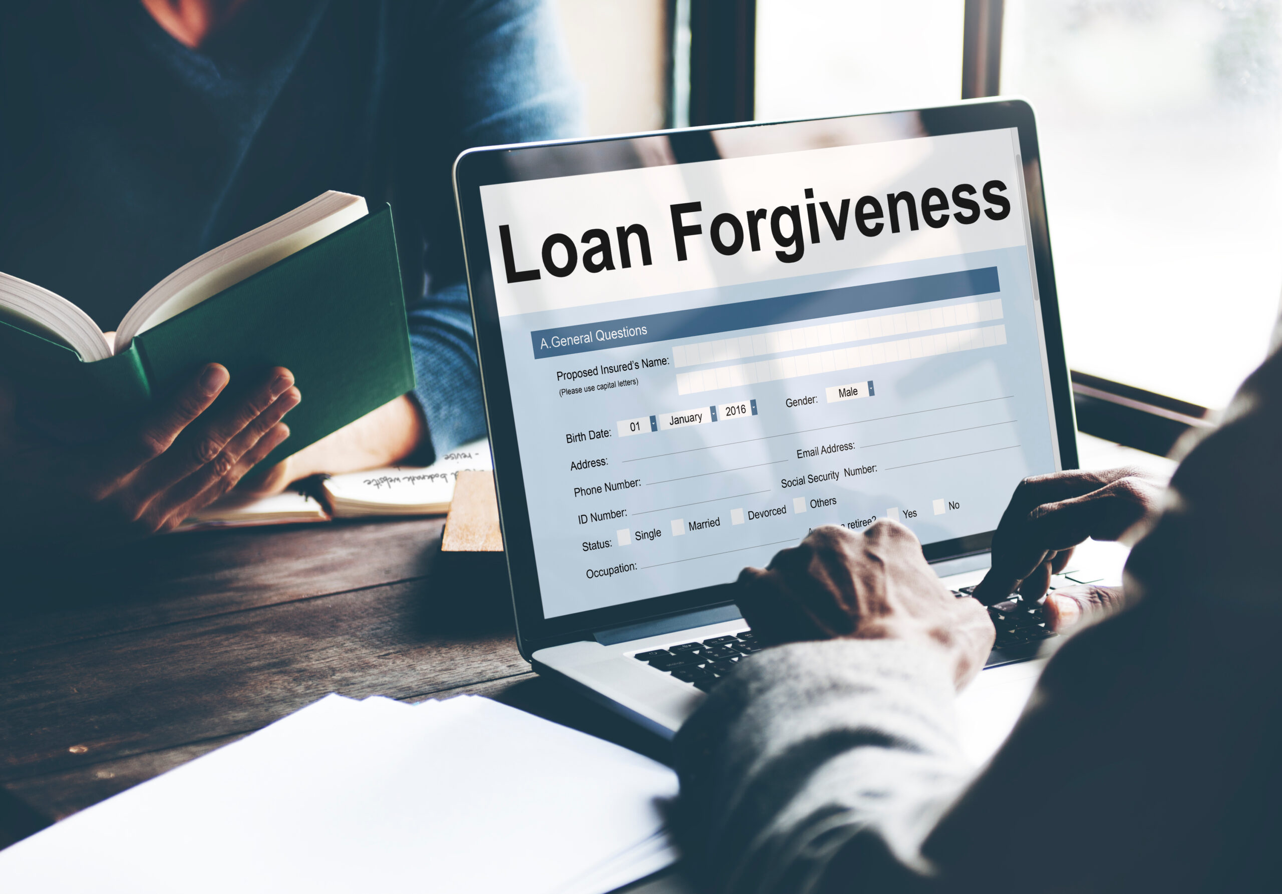 A person sits at an open laptop typing, LOAN FORGIVENESS is written across the top of the screen