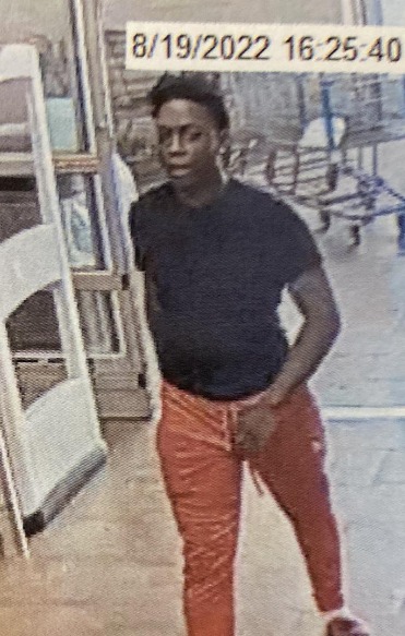 A Black male wearing orange pants and a black short sleeved shirt walks into a retail store in a still from security footage.