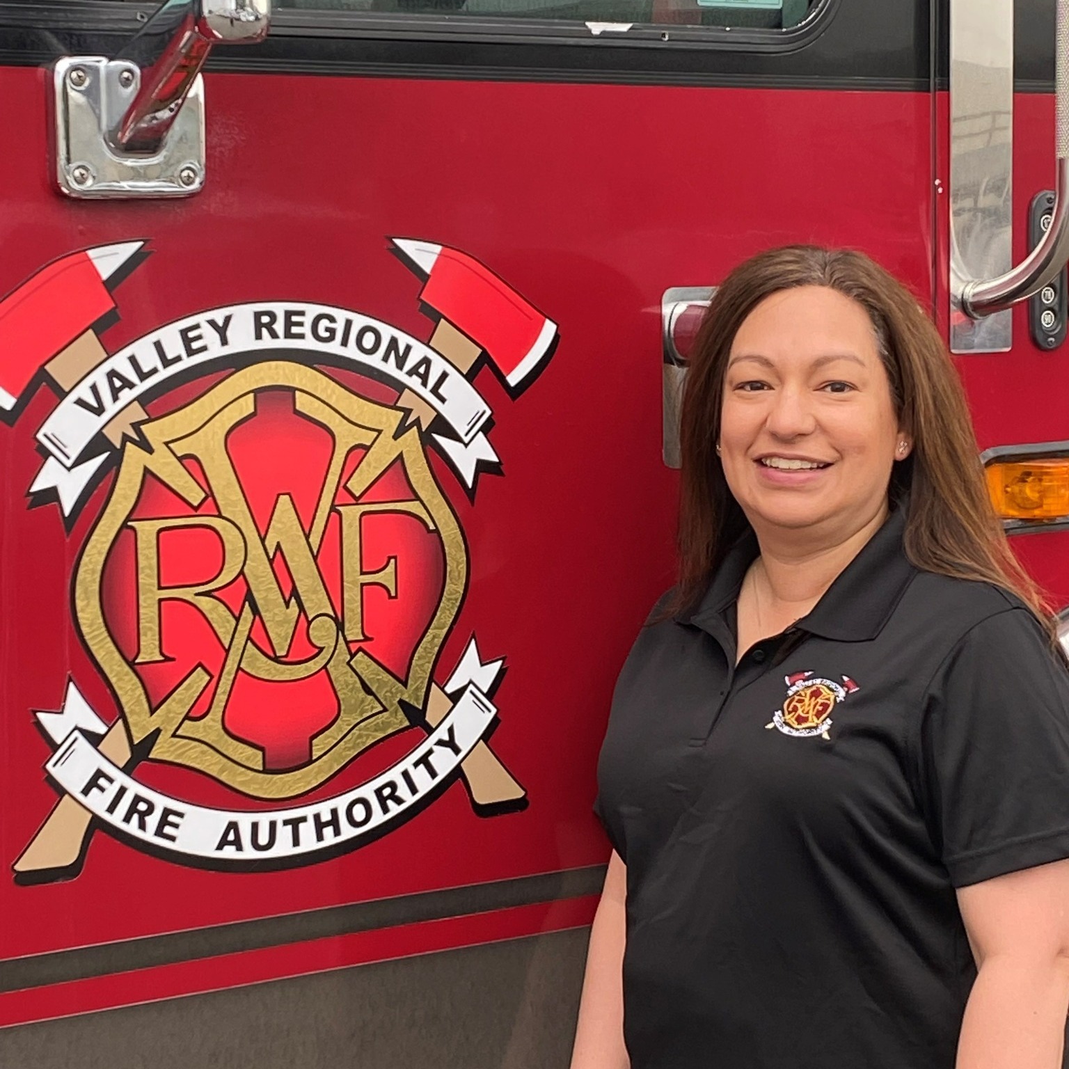 A mid-aged white female in a VRFA polo stands next to the VRFA logo on a red fire truck