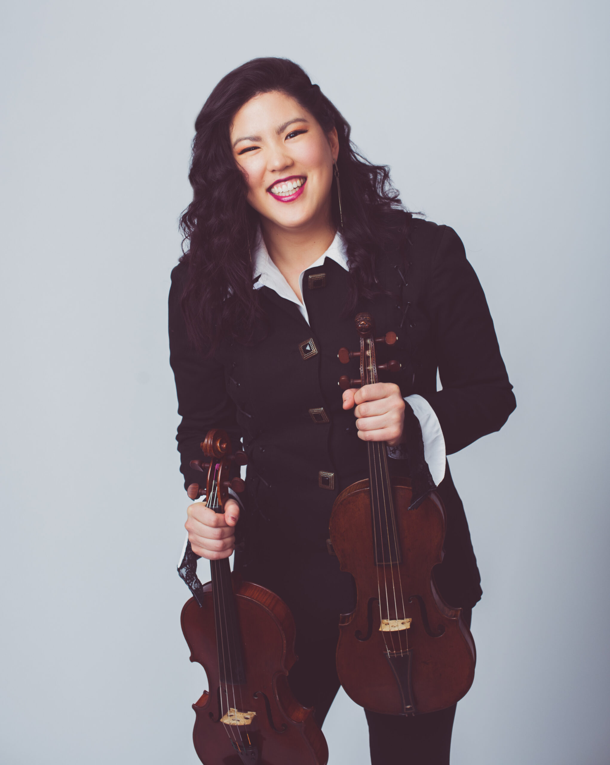 A young adult Asian female smiles, holding two violins. She wears a black button down top over a white shirt.