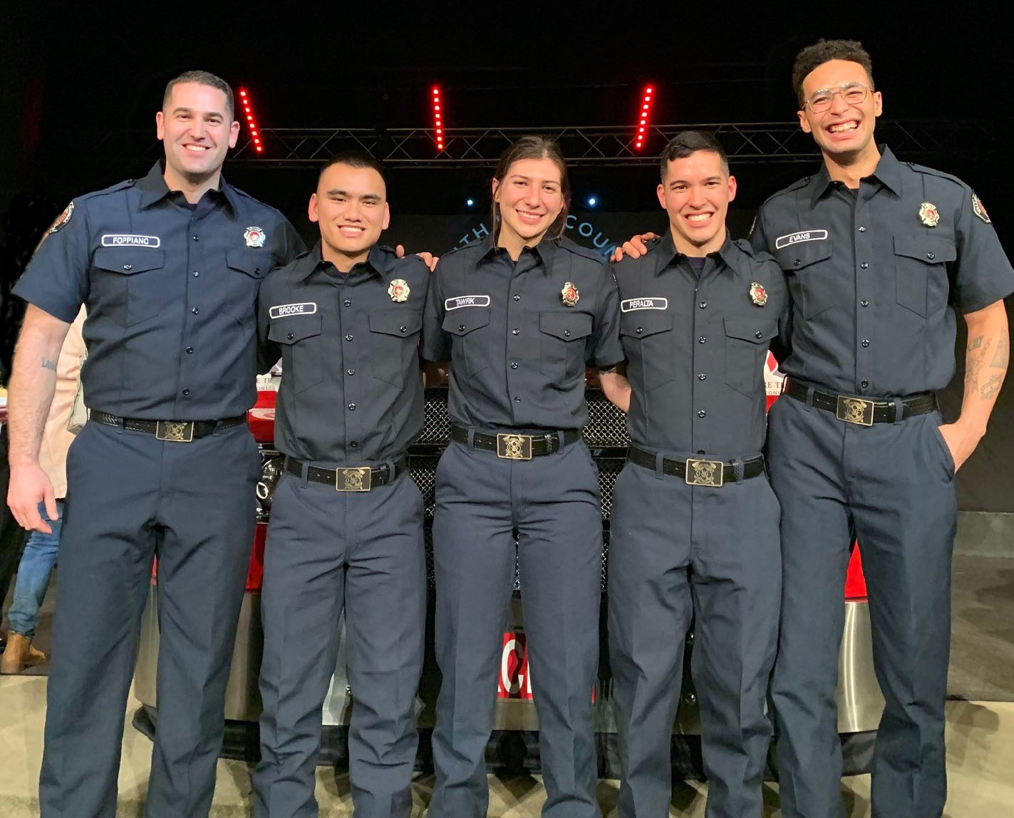 Five VRFA firefighters stand shoulder to shoulder in their dark blue uniforms, arms behind each others backs smiling. Two males flank a female on either side.