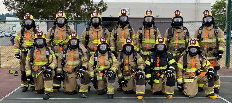 13 firefighters in turnout gear pose for a photo. The front line kneels with one knee up.