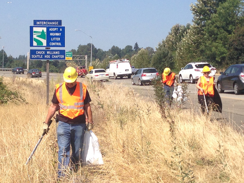 Threw members of a pick up crew wearing bright orange vests carrying trash pickers and trash bags as they collect litter along a Washington interstate
