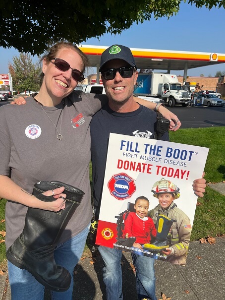A female (L) and male stand hugging shoulder to shoulder. The female holds a boot and the male holds a sign promoting fill the boot