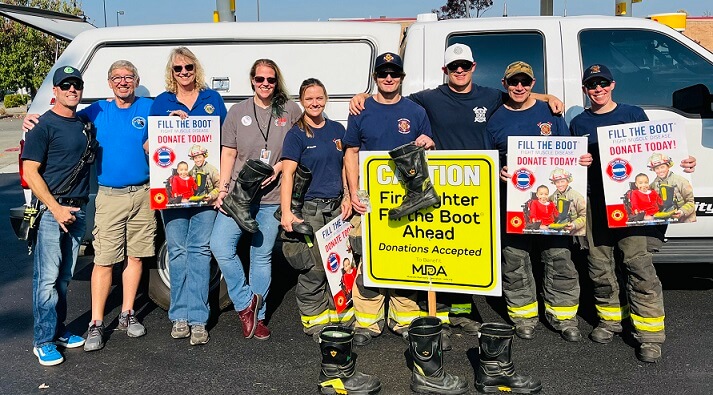 A group of firefighters and volunteers stand against a white pickup holding Fill the Boot signs and firefighter boots