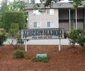 A wood sign painted white and green reading "Auburn Manor" set in a garden bed of mulch in front of an apartment complex.