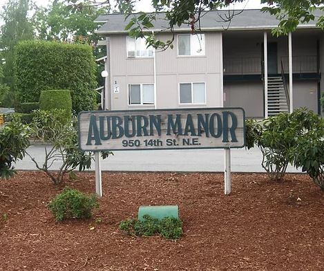 A wood sign painted white and green reading "Auburn Manor" set in a garden bed of mulch in front of an apartment complex.