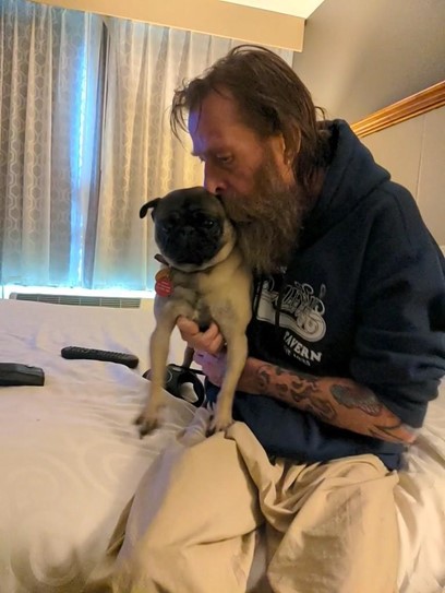 An older white male with medium length brown hair and beard sits on a bed holding a pug. The man hugs the dog close kissing its head. 